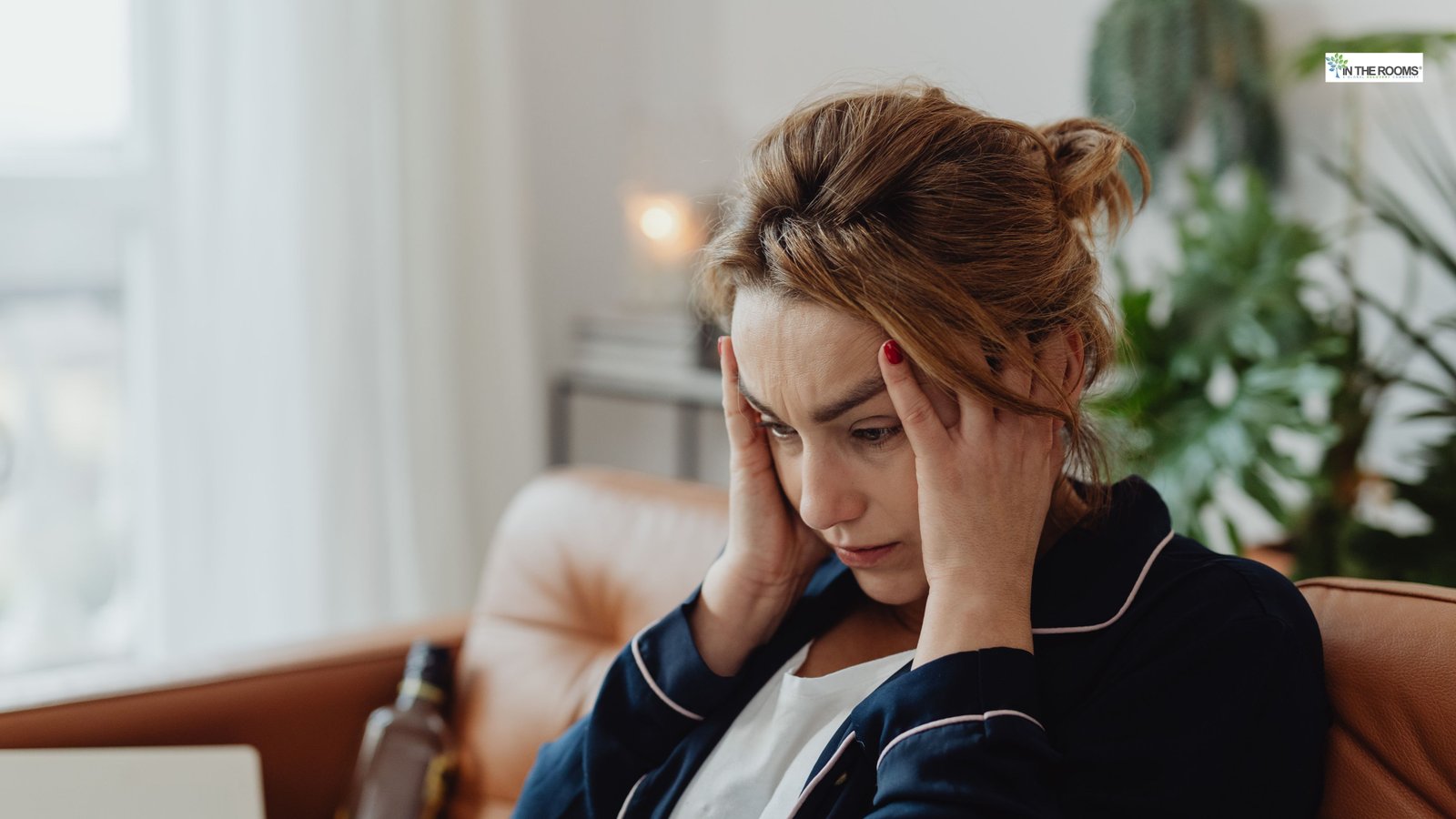 A woman sitting on a couch holding her head in distress, with a bottle visible nearby, representing the emotional impact of relapse.