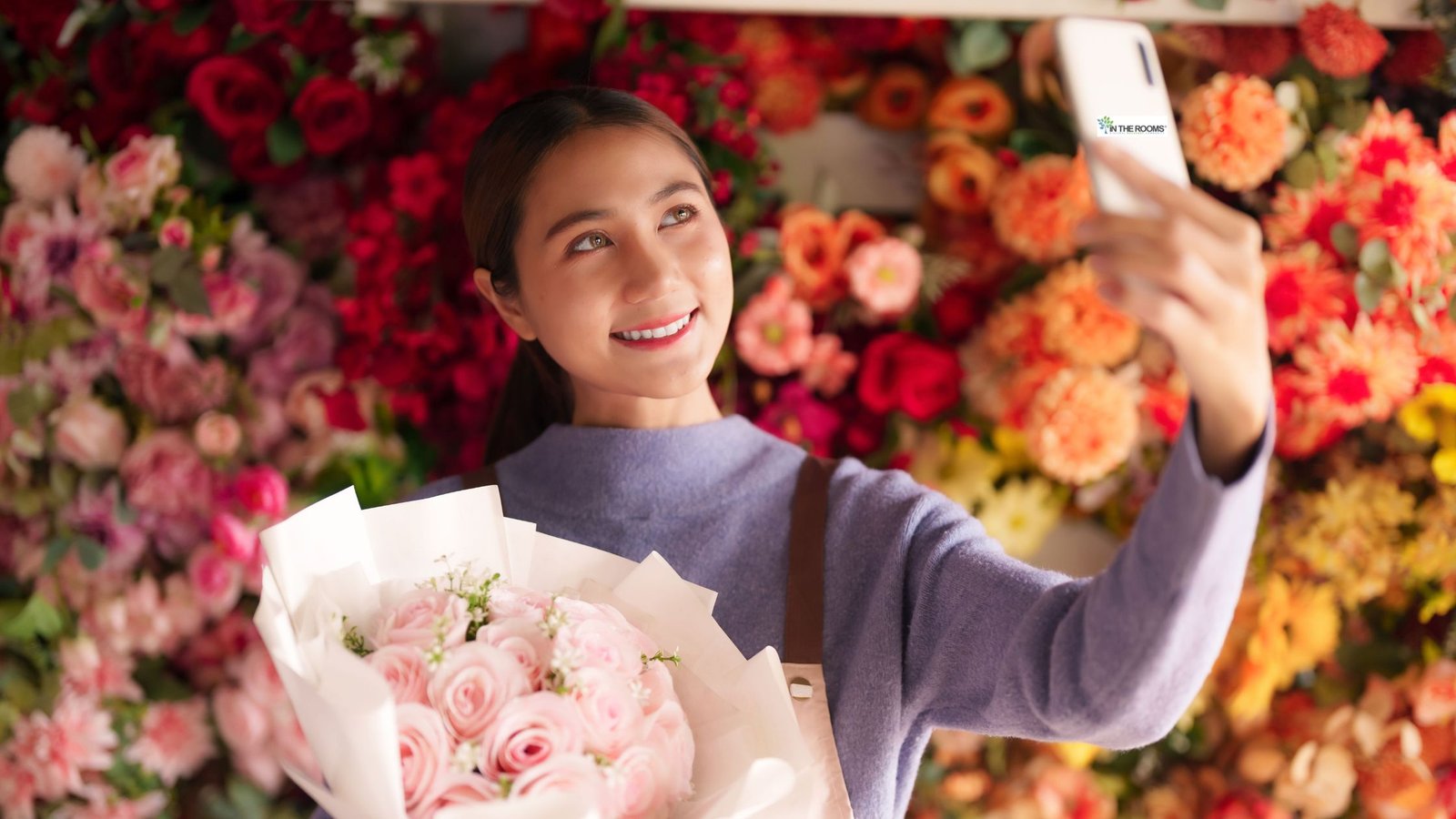 Woman smiling while taking a selfie and holding a bouquet of pink flowers in front of a colorful floral display.
