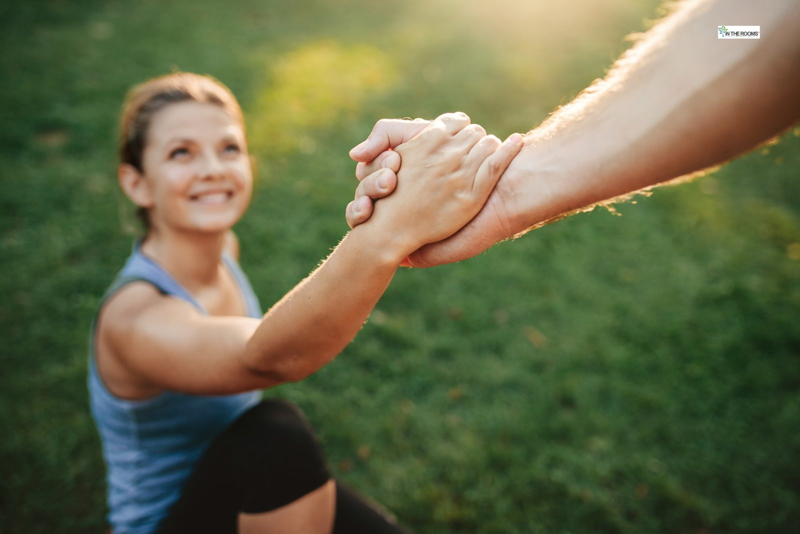 A smiling woman grasping another person’s hand as she is helped up outdoors, symbolizing support, trust, and reaching out for help.