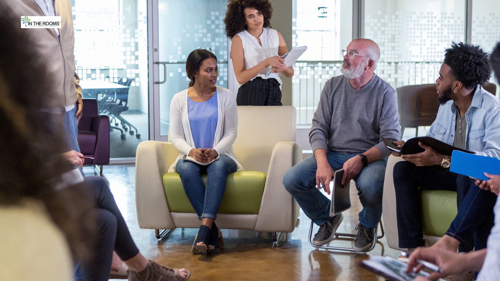 A diverse group of adults sitting in a circle during a community support group meeting, listening attentively to a woman standing and speaking.