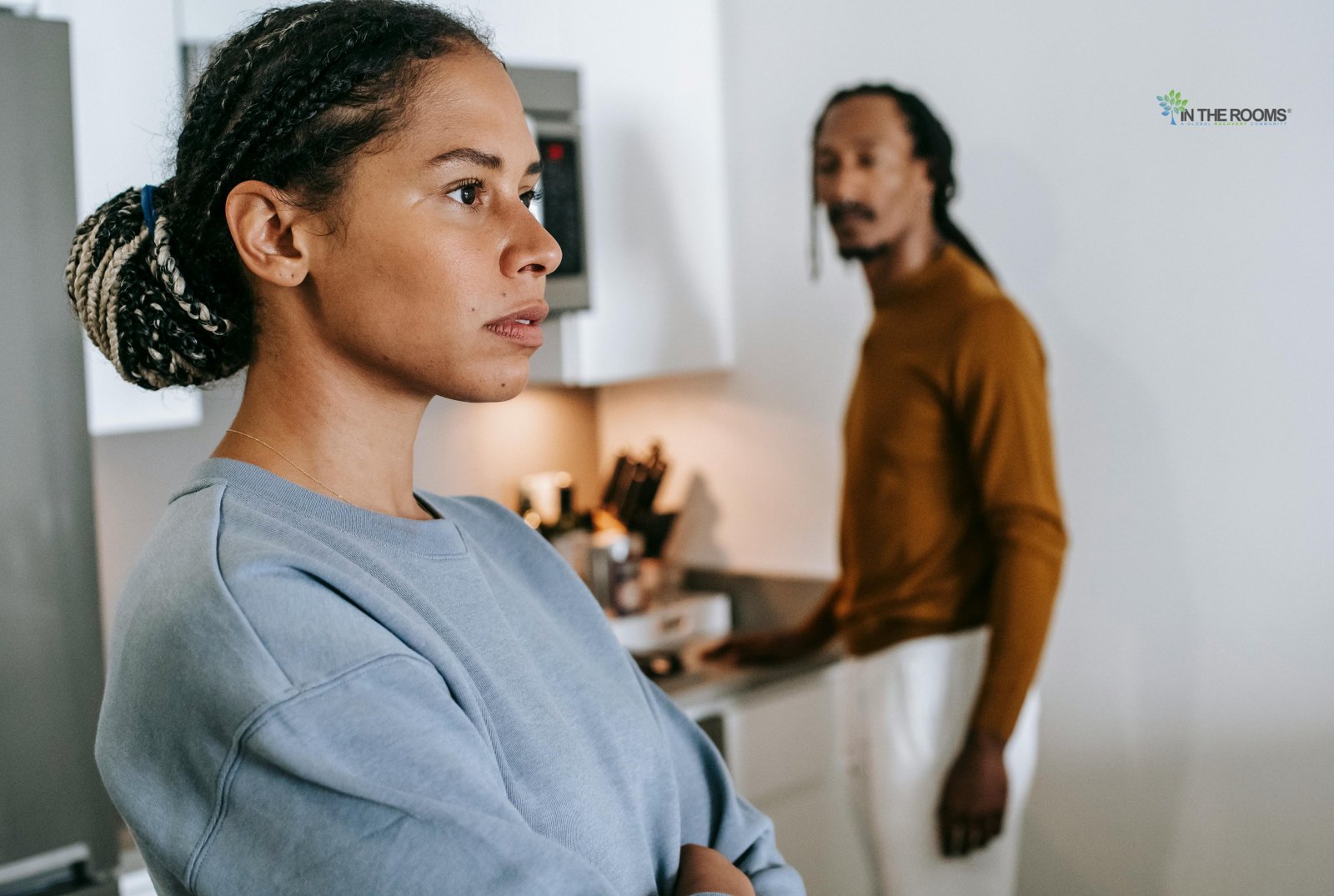 A concerned woman stands with folded arms while another person is visible in the background, illustrating the emotional challenges of supporting a loved one who is resistant to help.