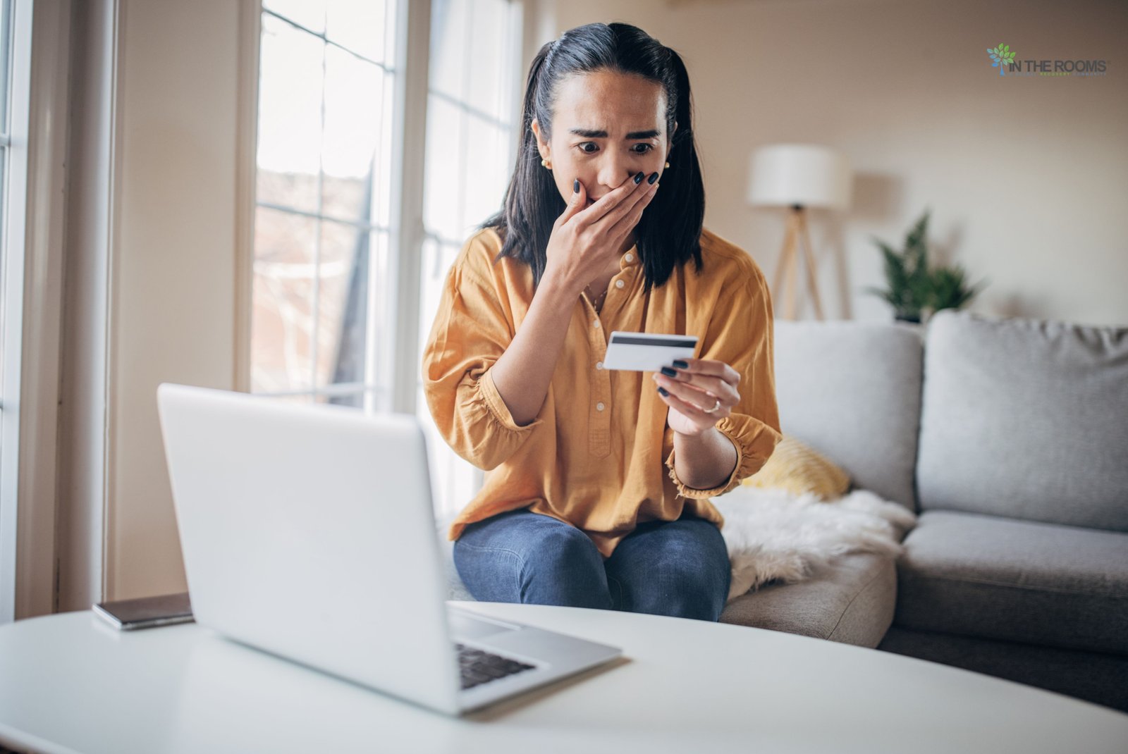 Person looking shocked while holding a credit card and using a laptop, highlighting online safety risks.