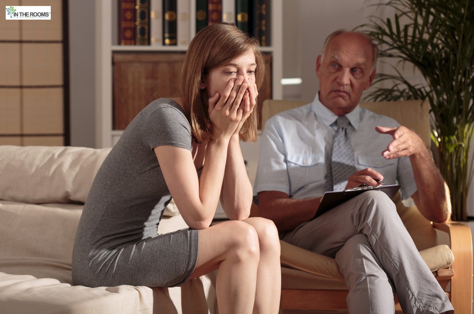 A woman sitting on a couch with her hands covering her mouth during a therapy session, while a therapist listens and takes notes.