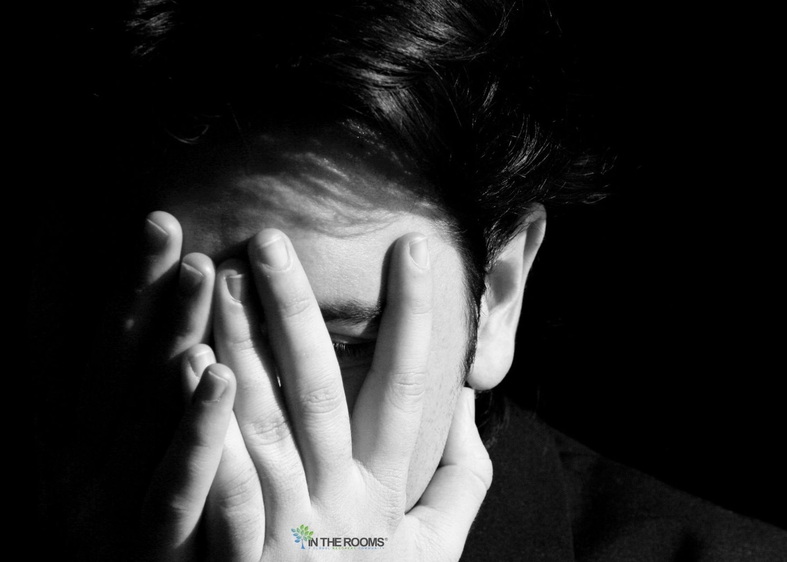 A person covers their face with their hands in a dark setting, symbolizing the shame, isolation, and emotional burden of stigma during recovery.