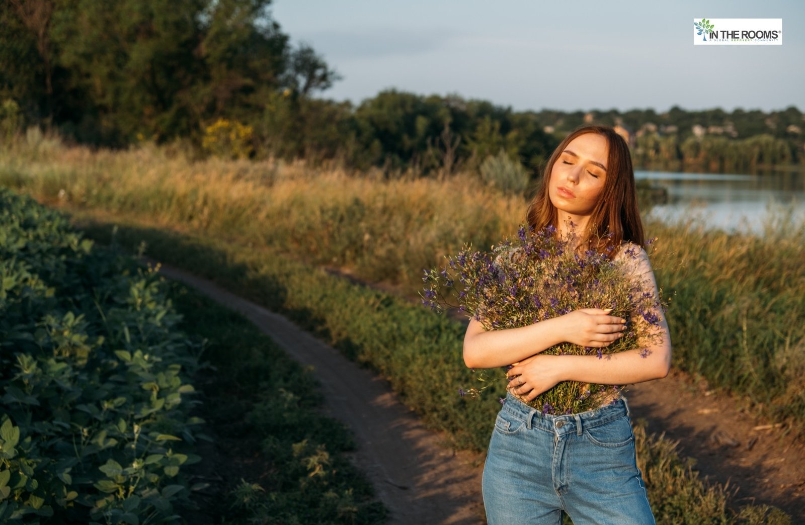 Woman standing outdoors on a dirt path, holding a bouquet of wildflowers with eyes closed, embracing peace and self-love.